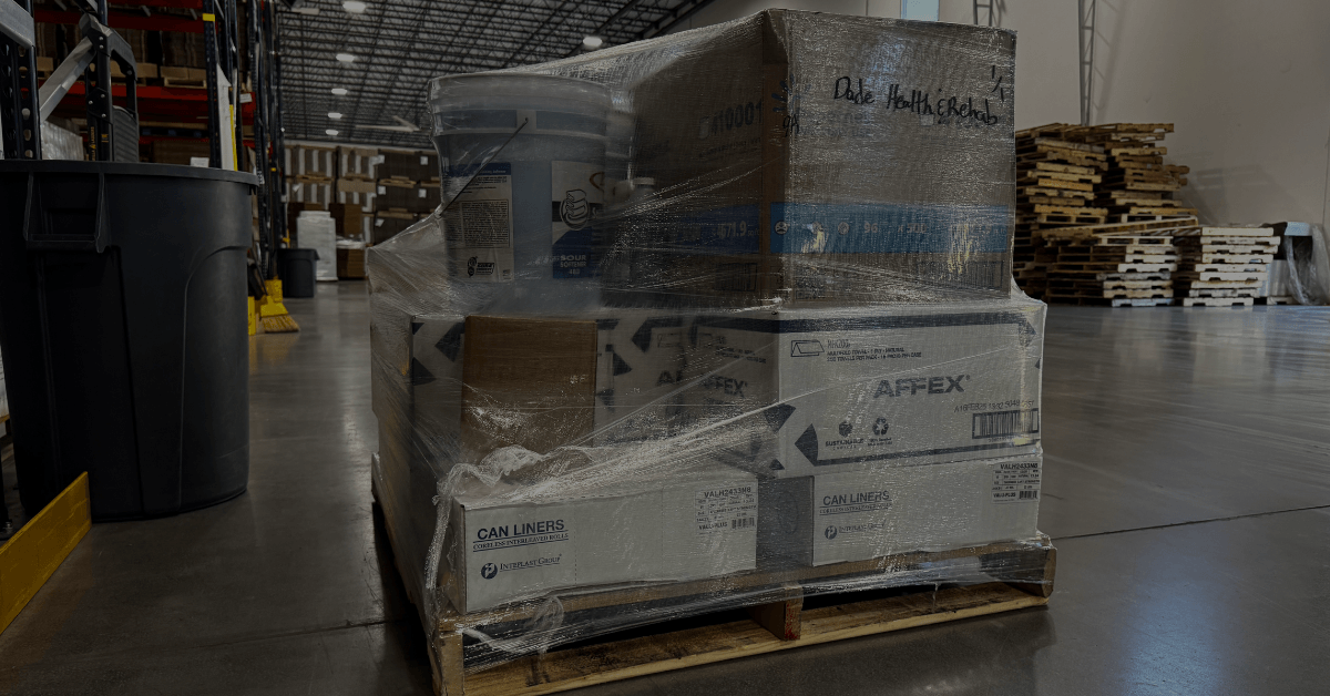 A wrapped pallet of boxed and jugged products near an open loading dock door, with a warehouse aisle visible behind it.