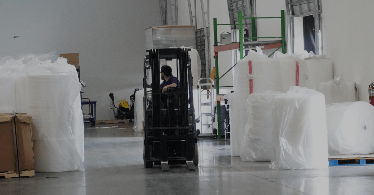 Chris Roberts driving a forklift through a warehouse aisle lined with large wrapped rolls of stretch film and pallet stock.